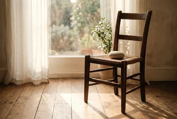 An empty wooden chair angled toward a bright window, warm low sun pooling on the floorboards, a single smooth stone resting on the seat, dust motes drifting in the still light.